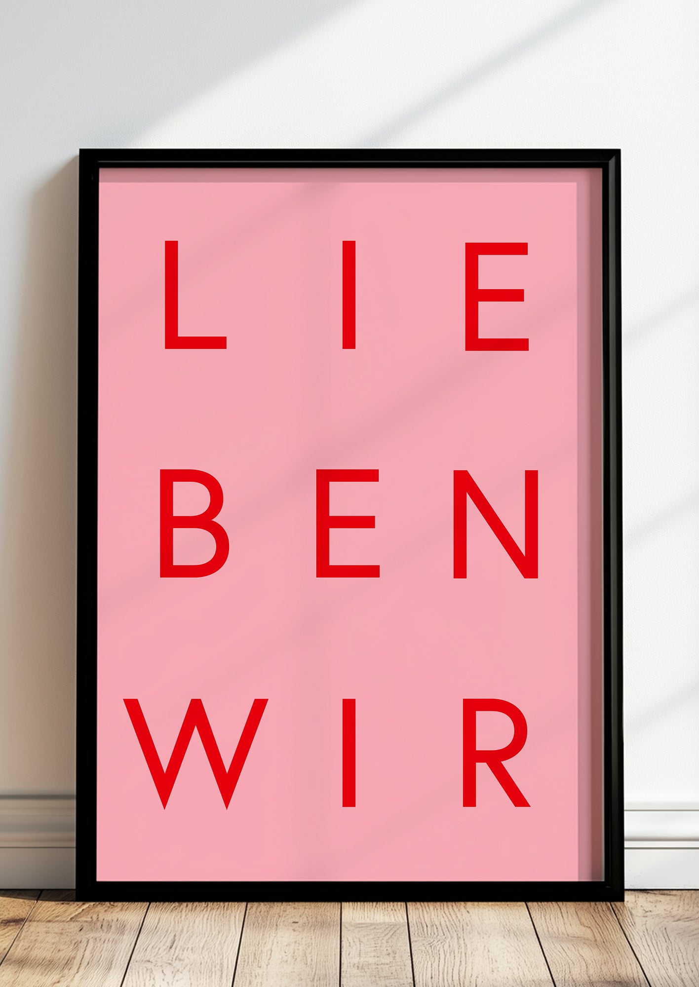 Rotes Typografieposter „LIEBEN WIR“ im schwarzen Holzrahmen, stehend auf dem Boden