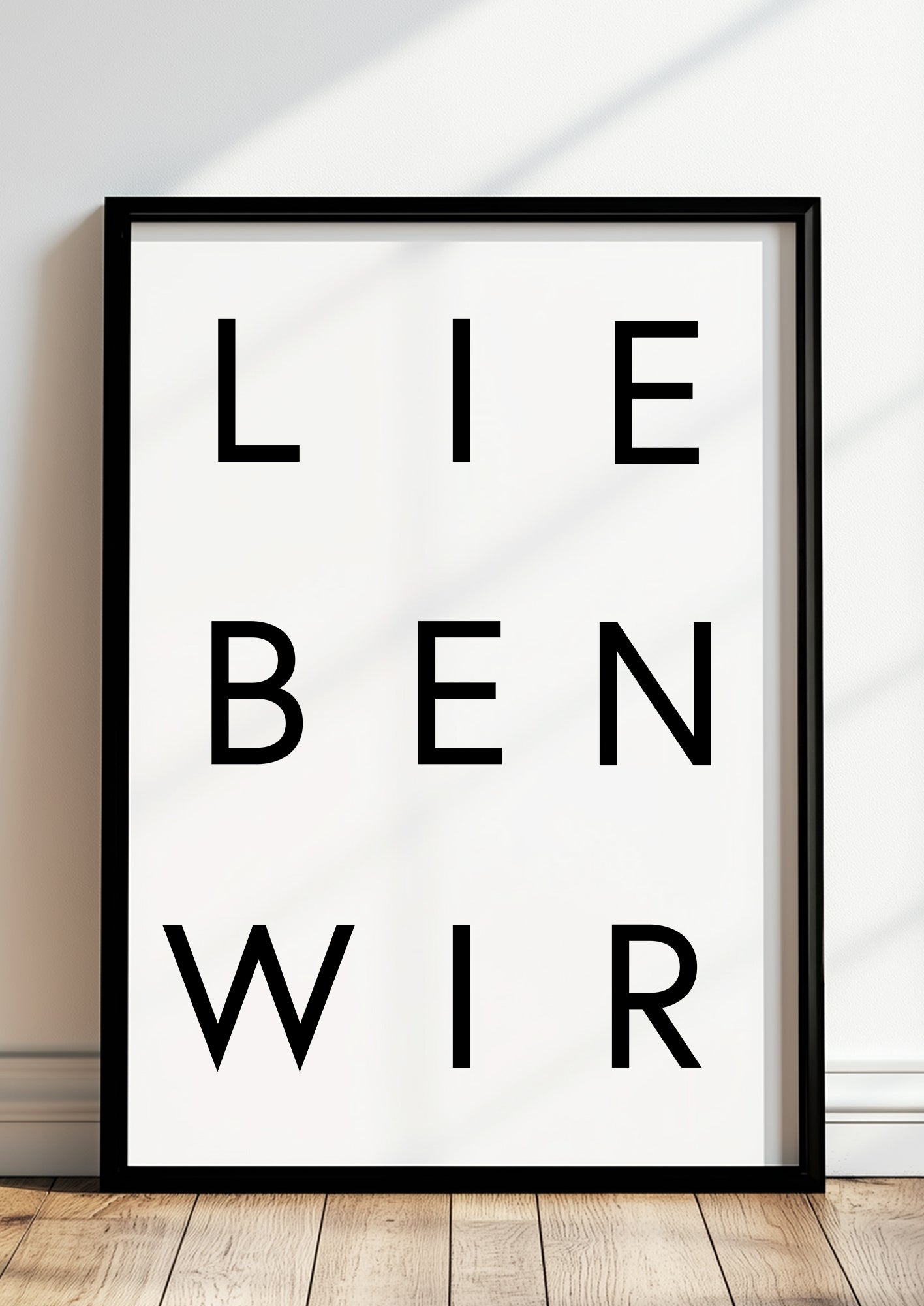 Schwarz-Weiß-Typografieposter „LIEBEN WIR“ im schwarzen Holzrahmen, stehend auf dem Boden