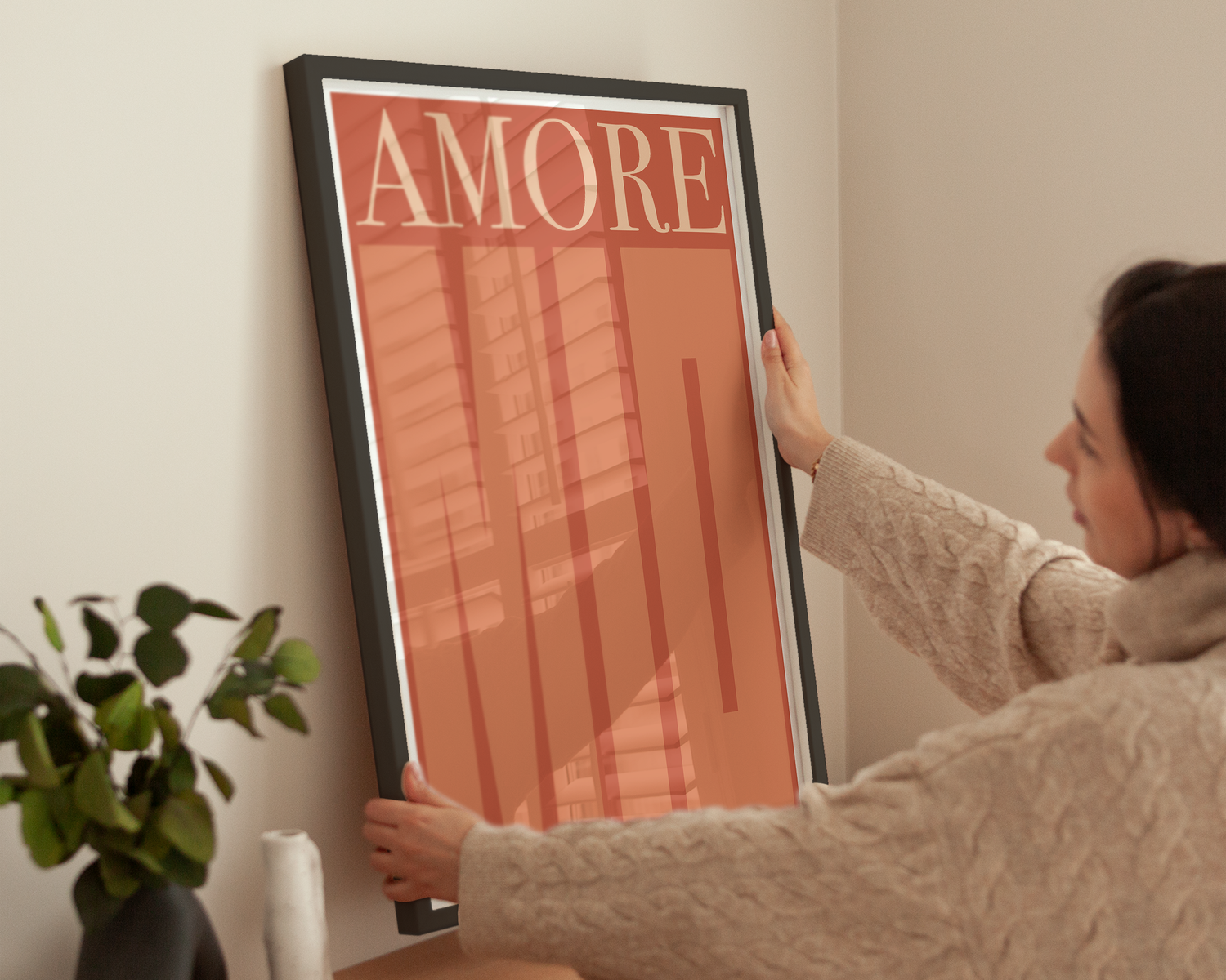 Frau richtet gerahmtes Kunstposter „Amore Mio“ an einer hellen Wand aus.
